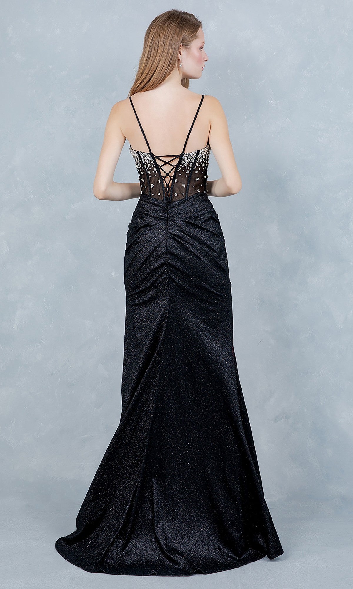 A woman stands facing away, wearing the Cinderella Couture 8562J—a long black prom dress featuring a jeweled corset bodice, lace-up back, thin straps, and a slightly ruched skirt.